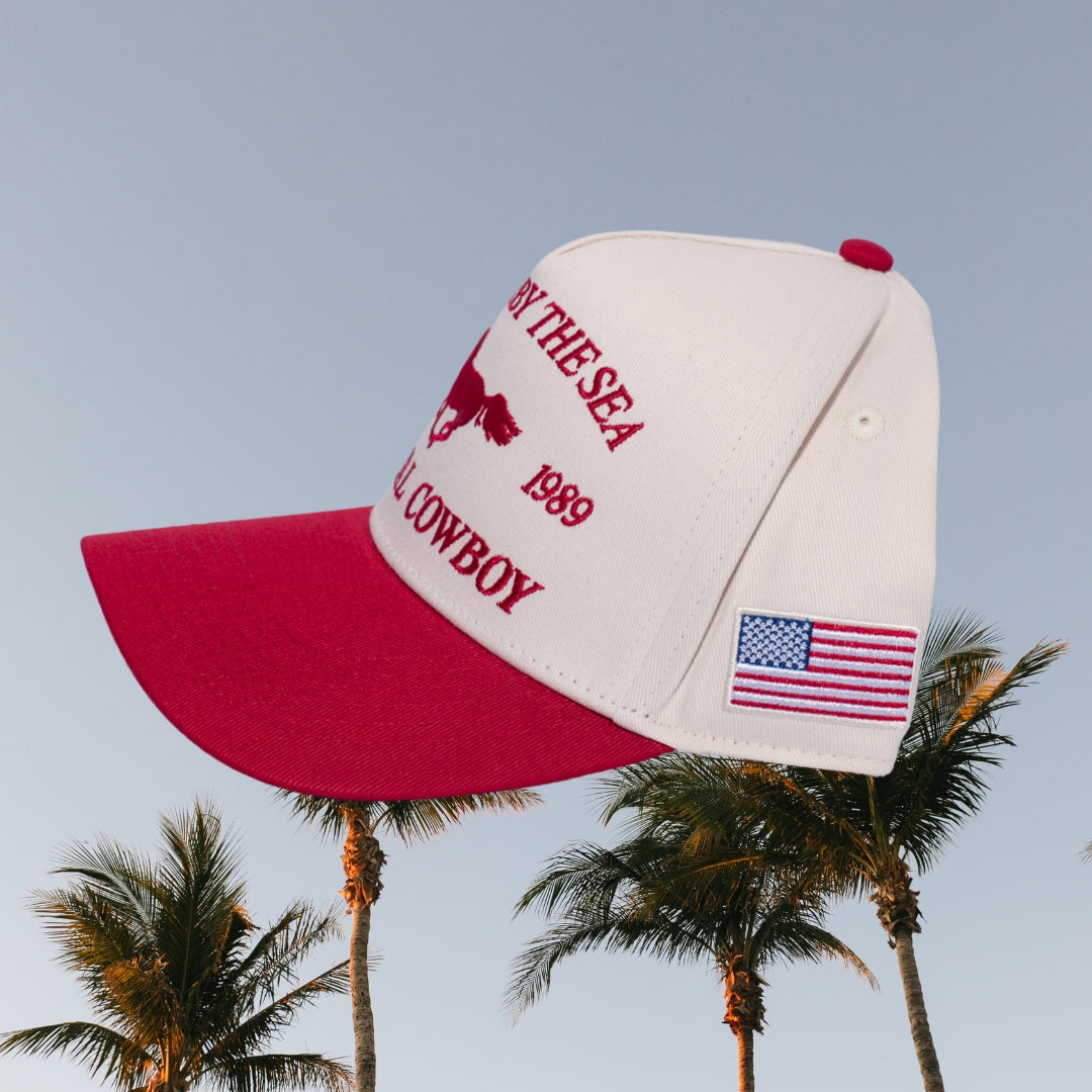 Side view of a white and red 5 panel trucker hat with 'CARMEL BY THE SEA CA 1989 COASTAL COWBOY' embroidery, an American flag logo, and a red horse on the front, set against palm tree background.