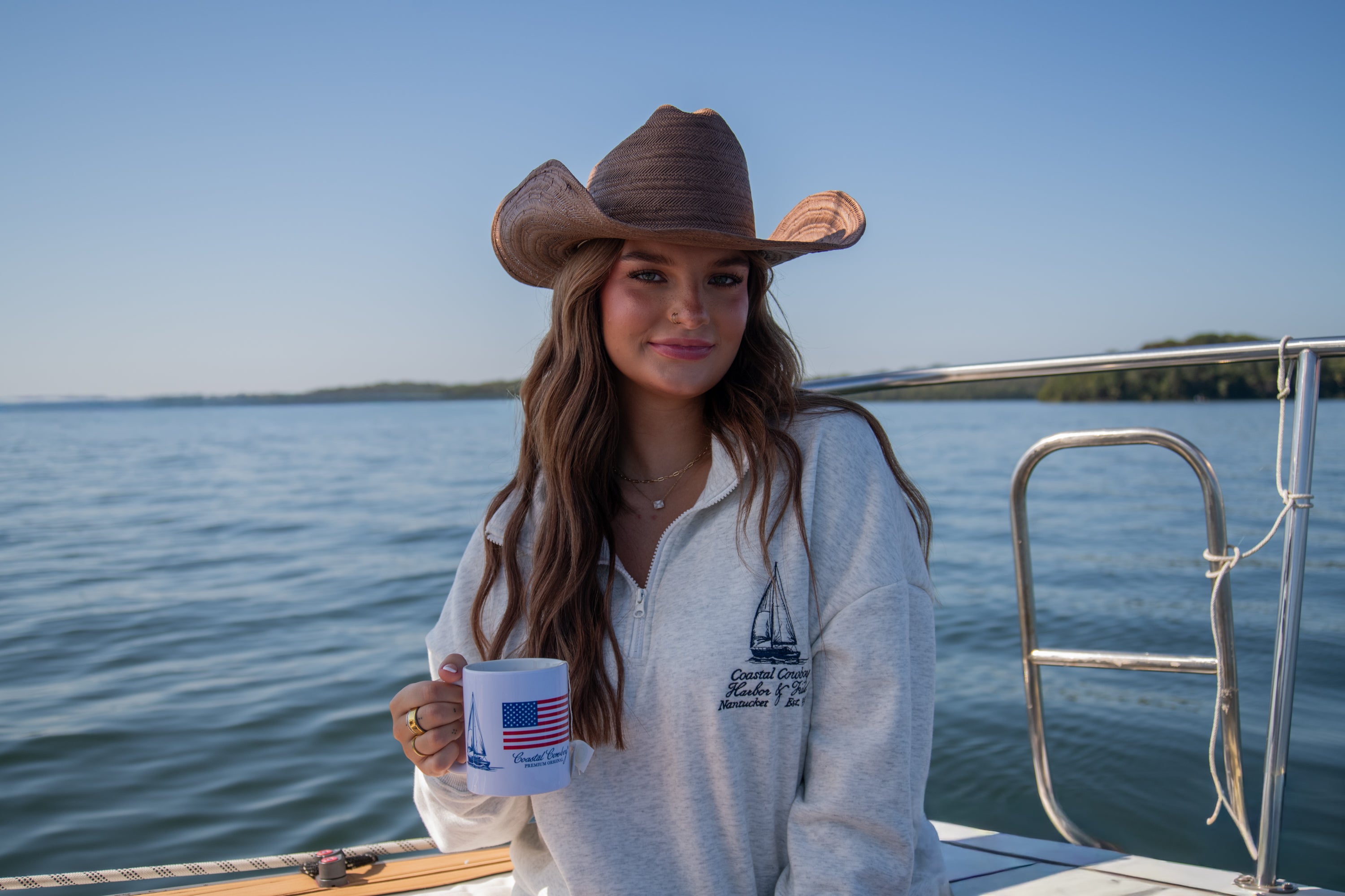 Woman wearing a cowboy hat and gray quarter zip with embroidered boat design on a boat holding a mug with a scenic background.
