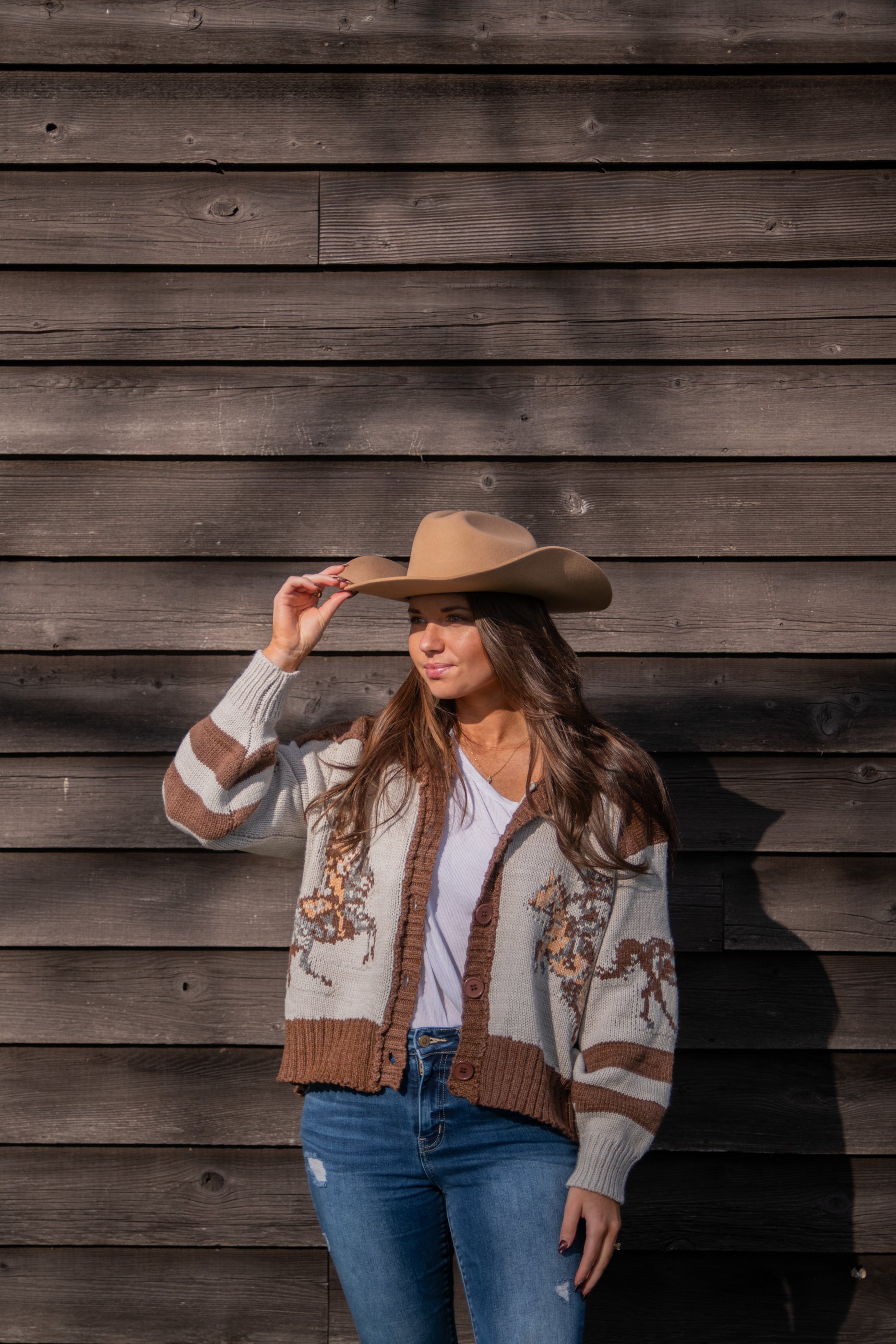 Woman wearing a patterned cardigan with horse design and jeans, holding a cowboy hat against a wooden wall.