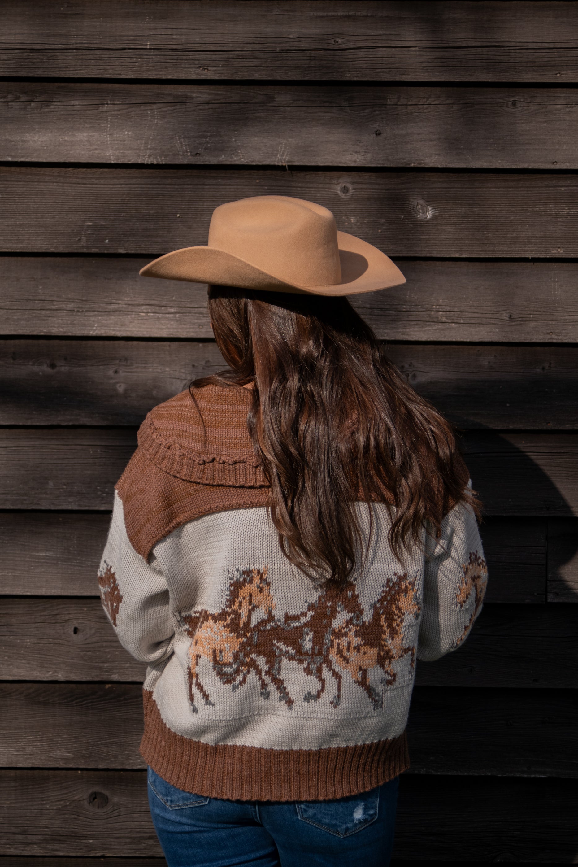 Person wearing a brown cowboy hat and a sweater with horse designs against a wooden wall.
