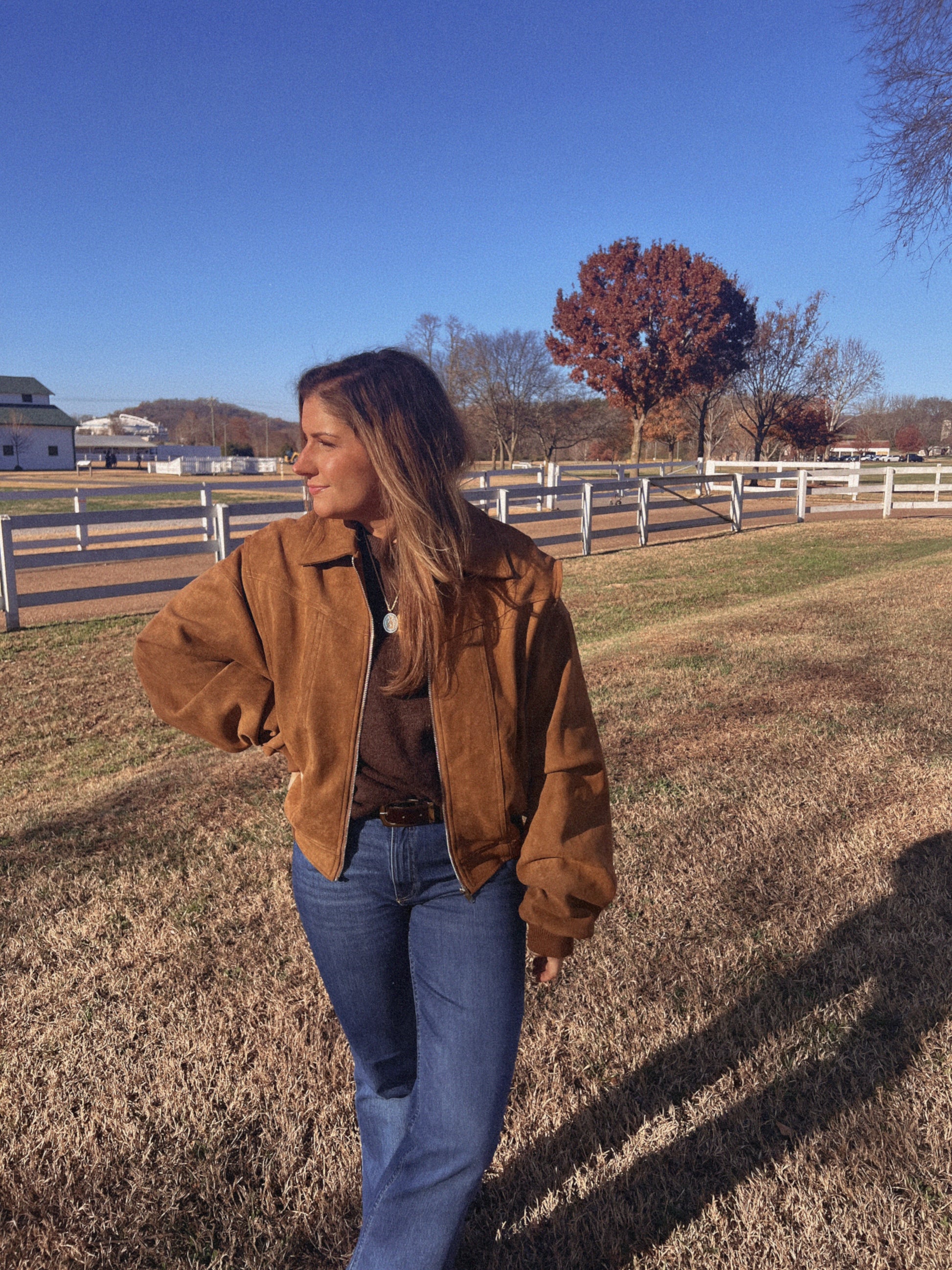 Person wearing a brown jacket and blue jeans standing outdoors with a clear blue sky.