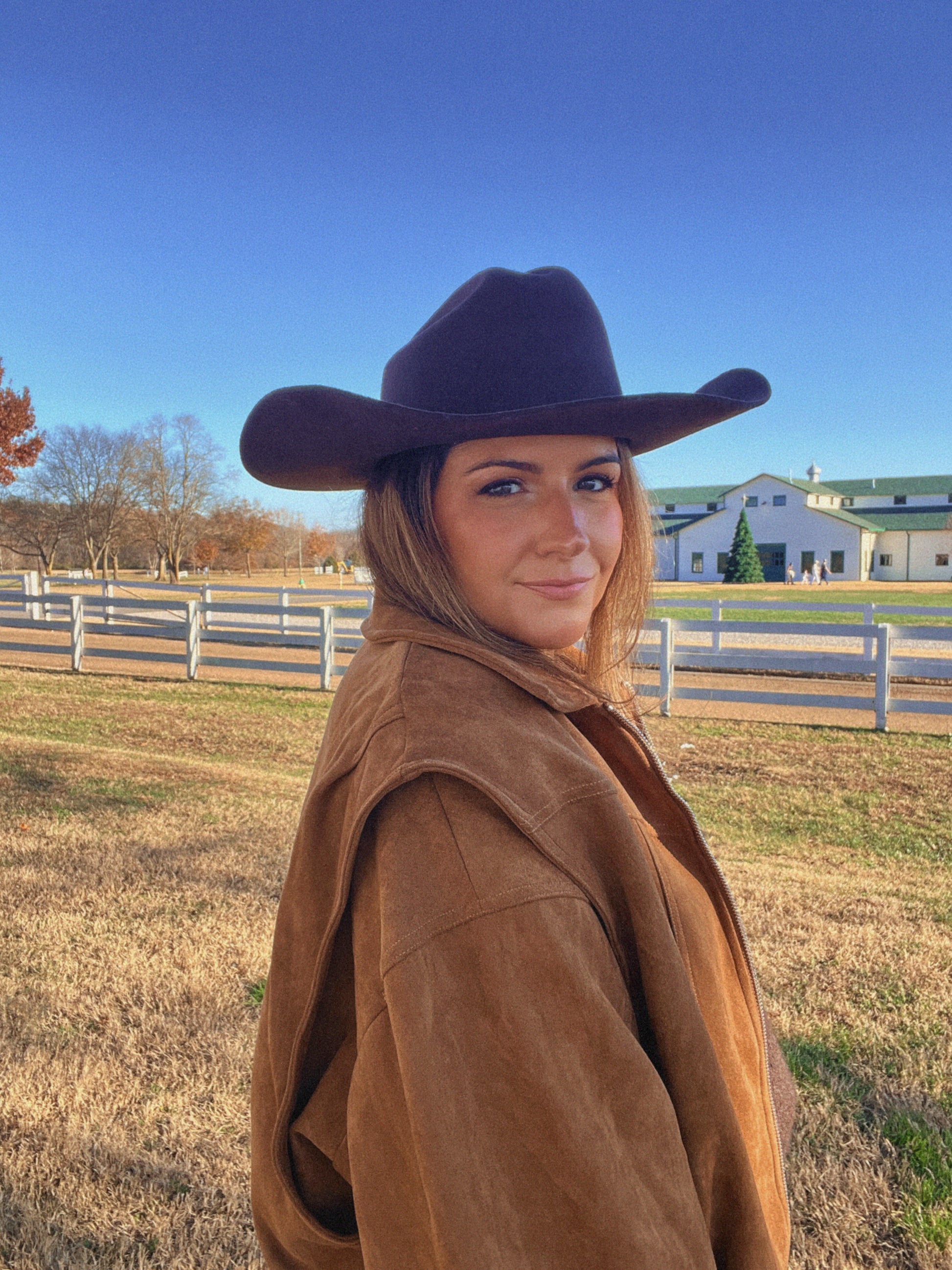 Person wearing a brown coat and cowboy hat in an outdoor setting with a fence and building in the background.